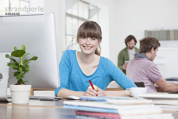 Porträt einer lächelnden jungen Frau  die an ihrem Schreibtisch in einem kreativen Büro sitzt.