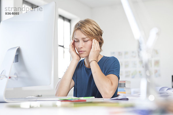 Porträt einer jungen Frau mit Kopfschmerzen am Schreibtisch in einem Kreativbüro