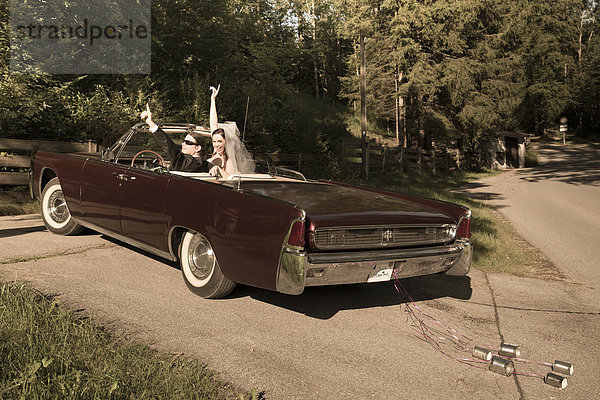 Brautpaar sitzend im Oldtimer-Cabriolet mit Siegeszeichen