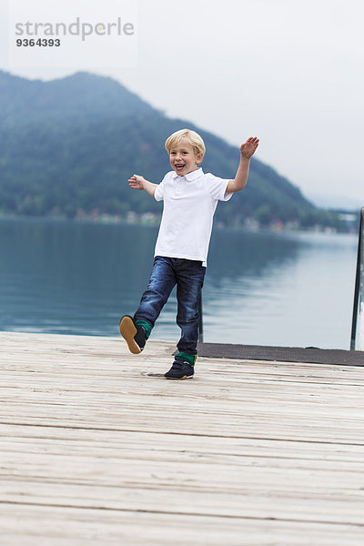 Lächelnder kleiner Junge  der auf einem Steg am See tanzt.