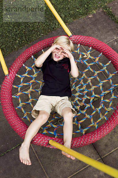 Porträt eines kleinen Jungen  der sich auf einem Spielplatz entspannt und seine Augen mit den Händen abschirmt.