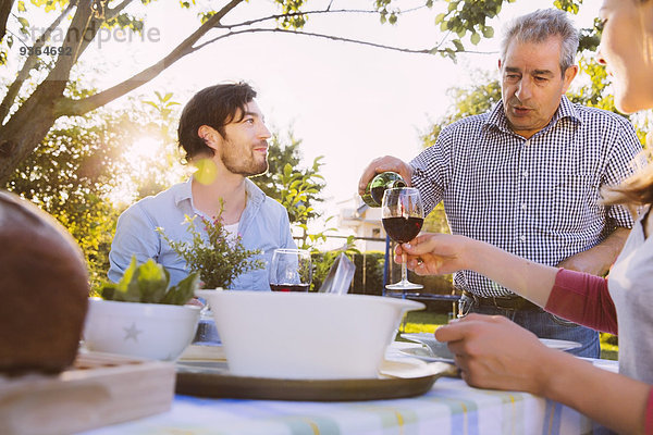 Senior Mann gießt Rotwein in Gläser eines Paares beim Abendessen im Garten.