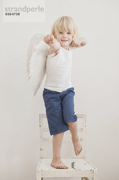 Porträt eines lächelnden kleinen Jungen mit abgewinkelten Flügeln  der auf einem Stuhl steht.