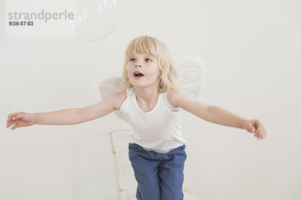 Porträt eines klaffenden kleinen Jungen mit angewinkelten Flügeln  der eine Seifenblase beobachtet.