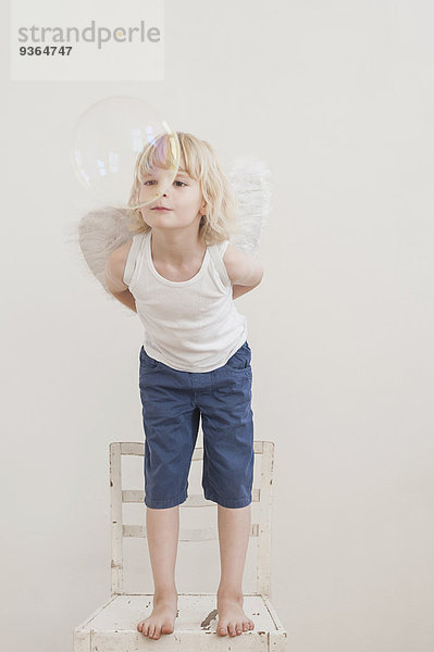 Porträt eines kleinen Jungen mit angewinkelten Flügeln  der eine Seifenblase beobachtet.