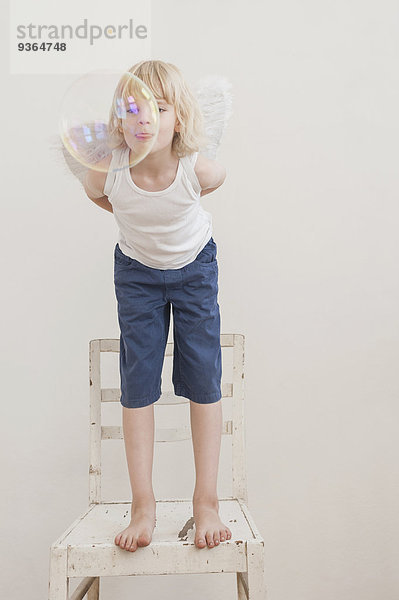 Porträt eines kleinen Jungen mit angewinkelten Flügeln  der eine Seifenblase beobachtet.