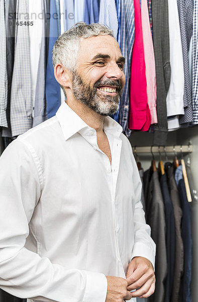 Porträt eines lächelnden Geschäftsmannes  der sein Hemd an seinem begehbaren Kleiderschrank zuknöpft.