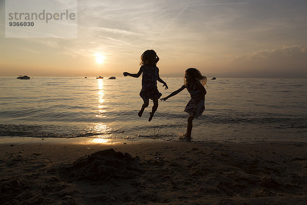 Italien  Gardasee  zwei Mädchen springen am Strand bei Sonnenuntergang