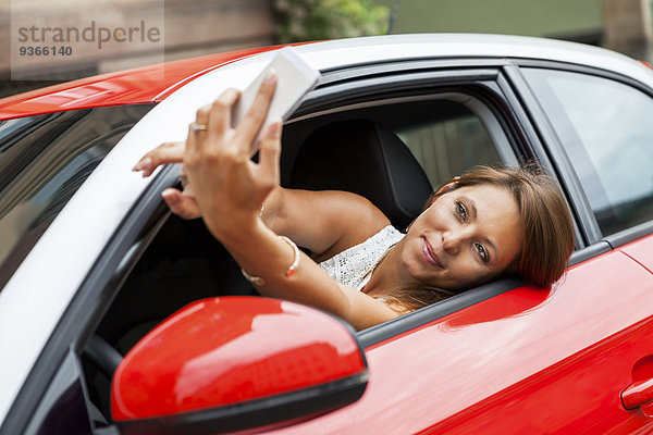 Porträt einer lächelnden jungen Frau  die in ihrem Auto sitzt und einen Selfie mit ihrem Smartphone nimmt.