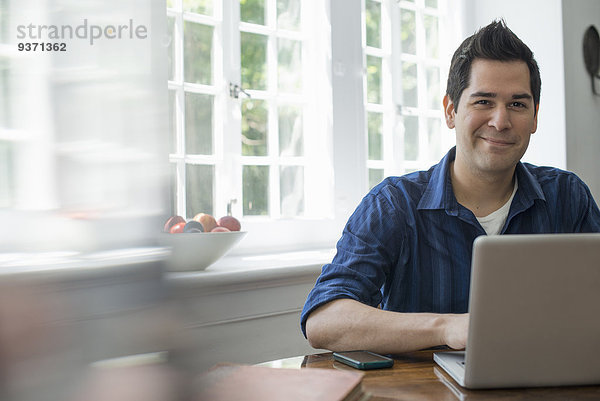 Ein Mann mit einem Laptop  der an einem Tisch am Fenster sitzt. Er arbeitet von zu Hause aus.