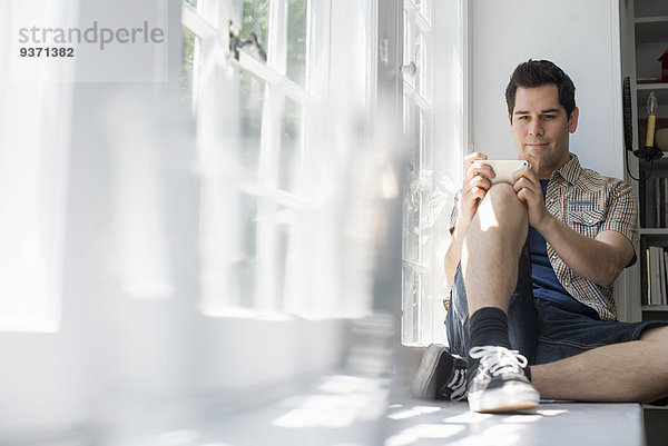 Am Fenster sitzender Mann mit einem Mobiltelefon in der Hand.