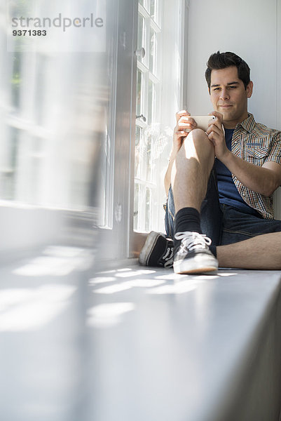 Am Fenster sitzender Mann mit einem Mobiltelefon in der Hand.