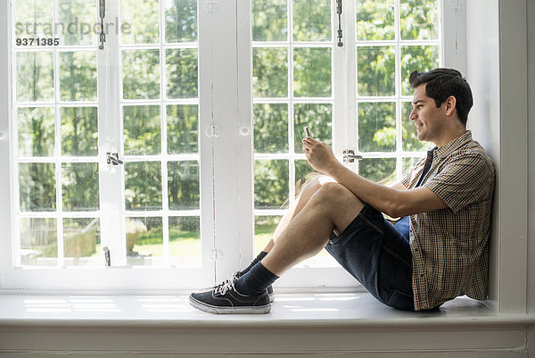 Am Fenster sitzender Mann mit einem Mobiltelefon in der Hand.