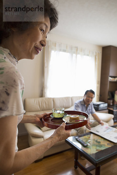 Eine Frau serviert einem Mann  der auf einem Sofa sitzt  ein Tablett mit Essen.