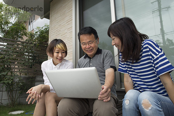 Ein Mann und zwei Frauen sitzen vor einem Haus. Sie halten einen Laptop-Computer in der Hand.