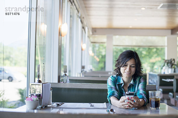 Eine Frau in kariertem Hemd  die lachend an einem Tisch sitzt und auf ihr Smartphone schaut.