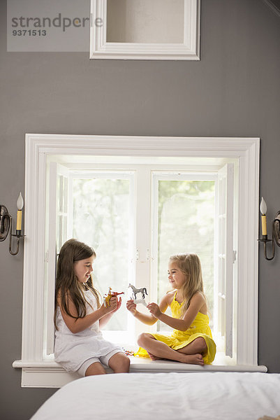 Zwei gemeinsam spielende Mädchen  die drinnen auf einem Fensterplatz sitzen.