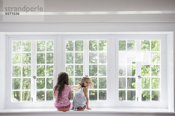 Zwei Mädchen sitzen nebeneinander an einem großen Fenster.