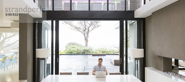 Junger Mann mit Laptop im modernen Esszimmer mit Terrassentüren und Swimmingpool im Hintergrund