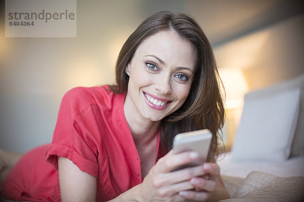 Porträt einer lächelnden Frau in rotem Kleid auf dem Bett liegend mit Smartphone