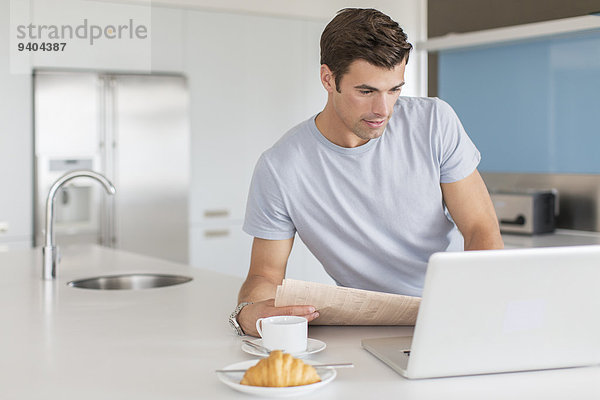 Mann mit Zeitung mit Laptop an der Küchenzeile  Kaffeetasse und Croissant im Vordergrund