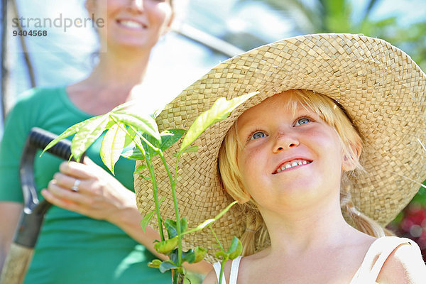 Mädchen mit Strohhut-Haltepflanze  Frau mit Schaufel im Hintergrund