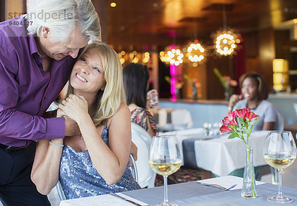 Lächelnder Mann umarmt Frau am Restauranttisch mit Weingläsern und Blumen
