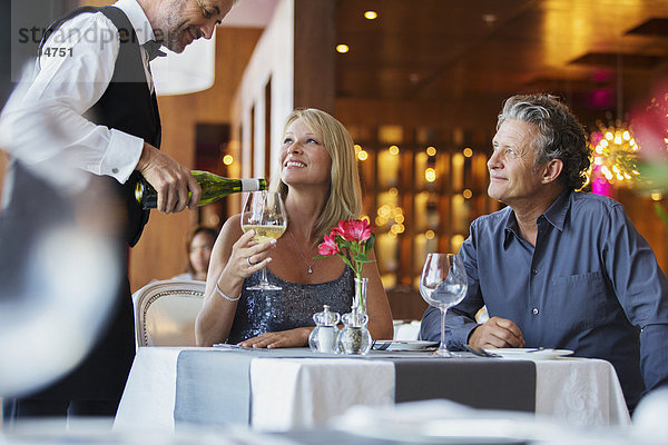 Ein reifes Paar sitzt am Restauranttisch  der Kellner gießt Weißwein in das Glas der Frau.