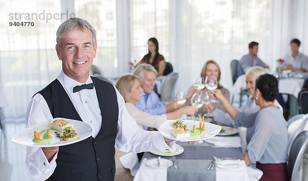 Porträt des Kellners  der den Teller mit ausgefallenen Gerichten hält  Menschen an Restauranttischen im Hintergrund