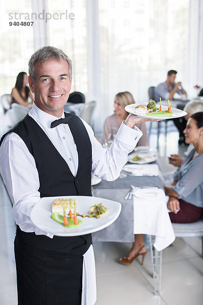 Porträt eines lächelnden Kellners mit ausgefallenen Gerichten  Menschen an Restauranttischen im Hintergrund