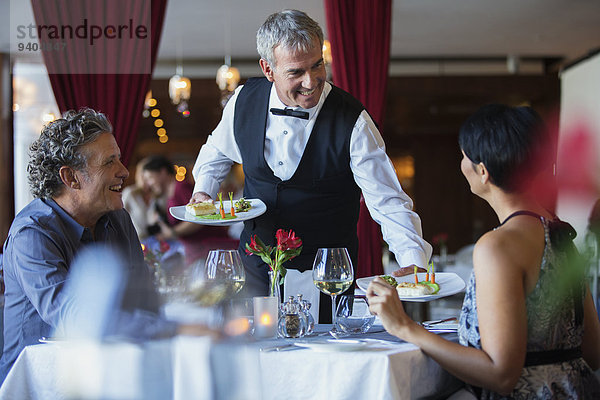 Lächelnder Kellner serviert ausgefallene Gerichte für ein reifes Paar  das am Tisch im Restaurant sitzt.