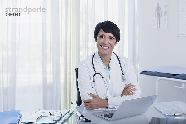 Porträt einer lächelnden Ärztin mit gekreuzten Armen am Schreibtisch mit Laptop