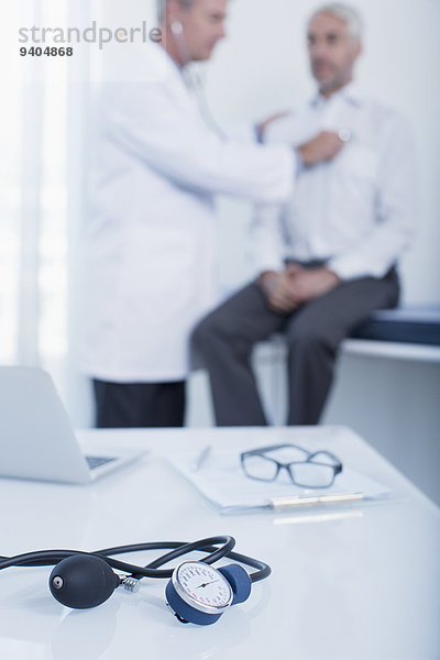 Blutdruckmessgerät  Laptop und Patientenkarte auf dem Schreibtisch in der Arztpraxis  Arzt untersucht Patient im Hintergrund