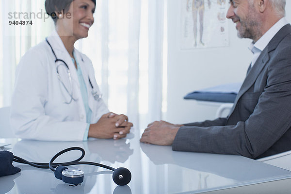 Lächelnde Ärztin im Gespräch mit Patientin am Schreibtisch im Büro  Blutdruckmessgerät im Vordergrund