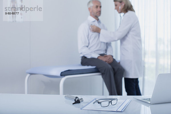 Ärztin untersucht Patientin im Büro  Laptop  Akte  Brille und Otoskop auf dem Schreibtisch im Vordergrund