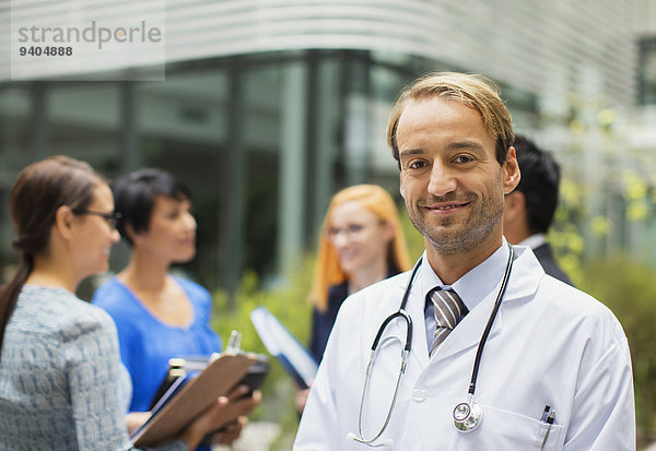 Porträt eines lächelnden Arztes im Laborkittel vor dem Krankenhaus  Frauen mit Klemmbrettern im Hintergrund