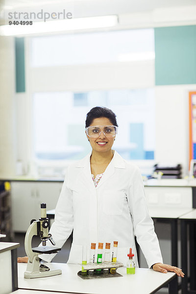 Porträt einer lächelnden Lehrerin mit Schutzbrille  die hinter dem Schreibtisch mit Mikroskop und Reagenzgläsern im Rack steht.