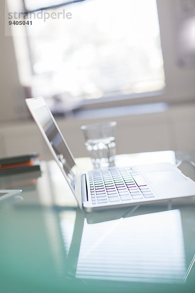 Offener Laptop und Wasserglas auf Glasschreibtisch im Büro