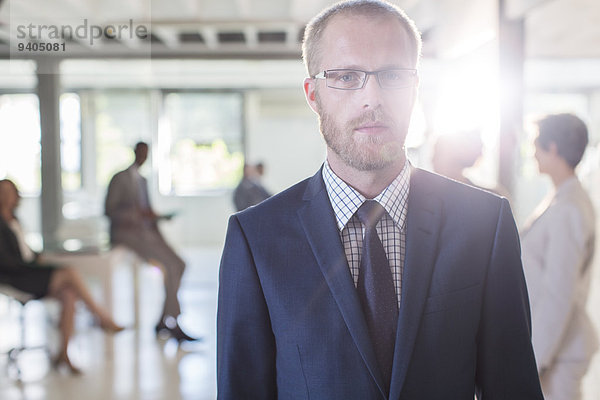 Porträt eines seriösen Geschäftsmannes mit Brille und Anzug im Büro  Kollegen im Hintergrund