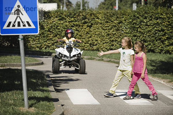 Two girls walking on zebra crossing with boy on quadbike waiting