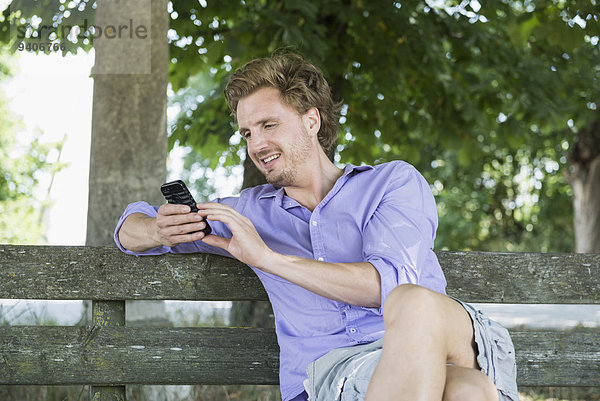 sitzend benutzen Mann lächeln Sitzbank Bank Mittelpunkt Smartphone Erwachsener