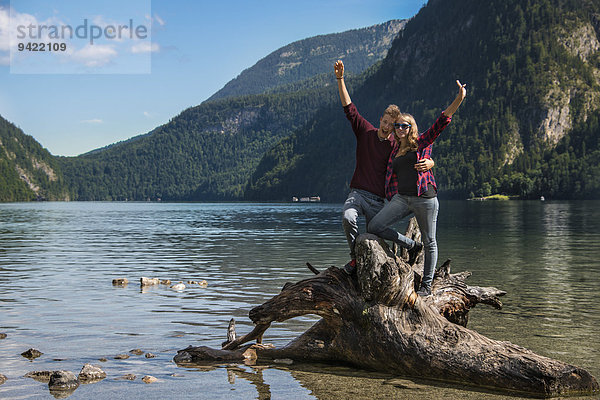 Junges Pärchen auf einem umgefallenen Baumstamm strecken Arme in die Luft  Königssee  Nationalpark Berchtesgaden  Berchtesgadener Land  Oberbayern  Bayern  Deutschland  Europa