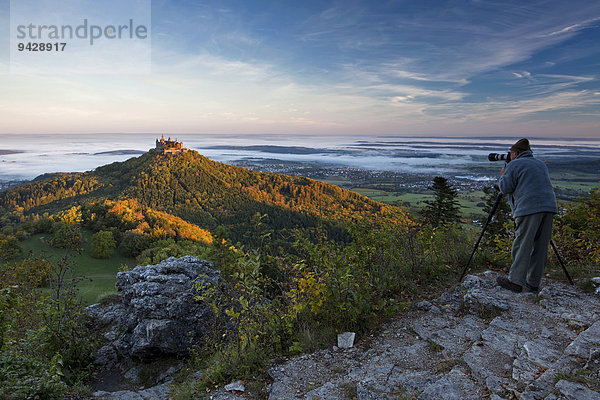 Die Burg Hohenzollern mit Fotograf im morgendlichen Herbstlicht mit Herbstwald  Frühnebel. Schwäbische Alb  Deutschland  Europa