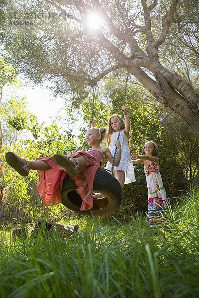 Niederwinkelansicht von drei Mädchen  die auf einer Baumreifenschaukel im Garten spielen.