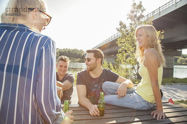 Vier junge Freunde trinken Bier auf der Picknickbank am Flussufer