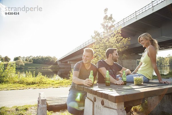 Drei junge Freunde trinken Bier auf der Picknickbank am Flussufer