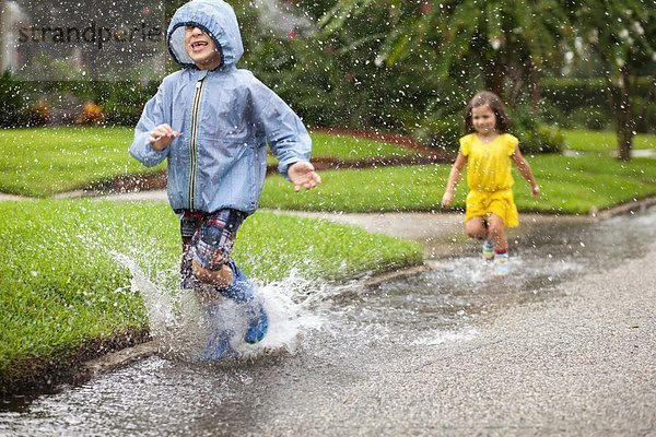 Junge und Schwester in Gummistiefeln laufen und planschen in der Regenpfütze