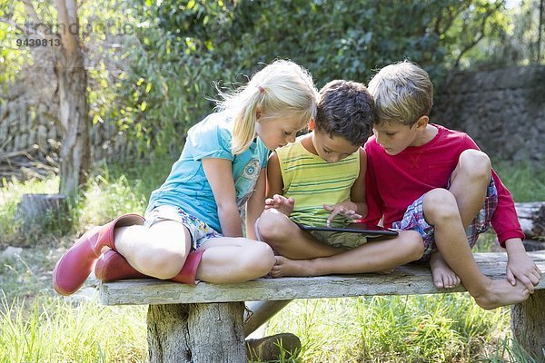 Drei Kinder sitzen auf einem Gartensitz und schauen auf ein digitales Tablett.