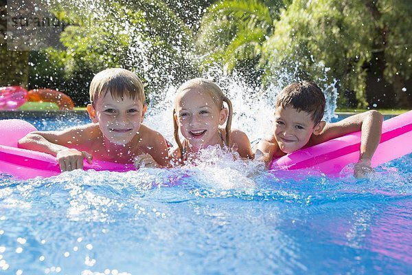 Porträt von drei Kindern  die auf einer aufblasbaren Matratze im Gartenschwimmbad planschen.