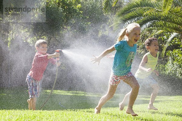 Drei Kinder im Garten jagen sich gegenseitig mit Wassersprenger